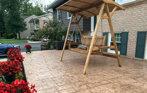 concrete-patios Wooden porch swing with canopy on a stamped concrete patio, red flowers in black pots, houses in background.