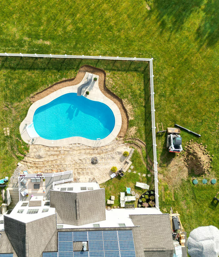 project-01 Aerial view of a backyard with a kidney-shaped pool, patio, and lawn, partially fenced, with construction equipment nearby.