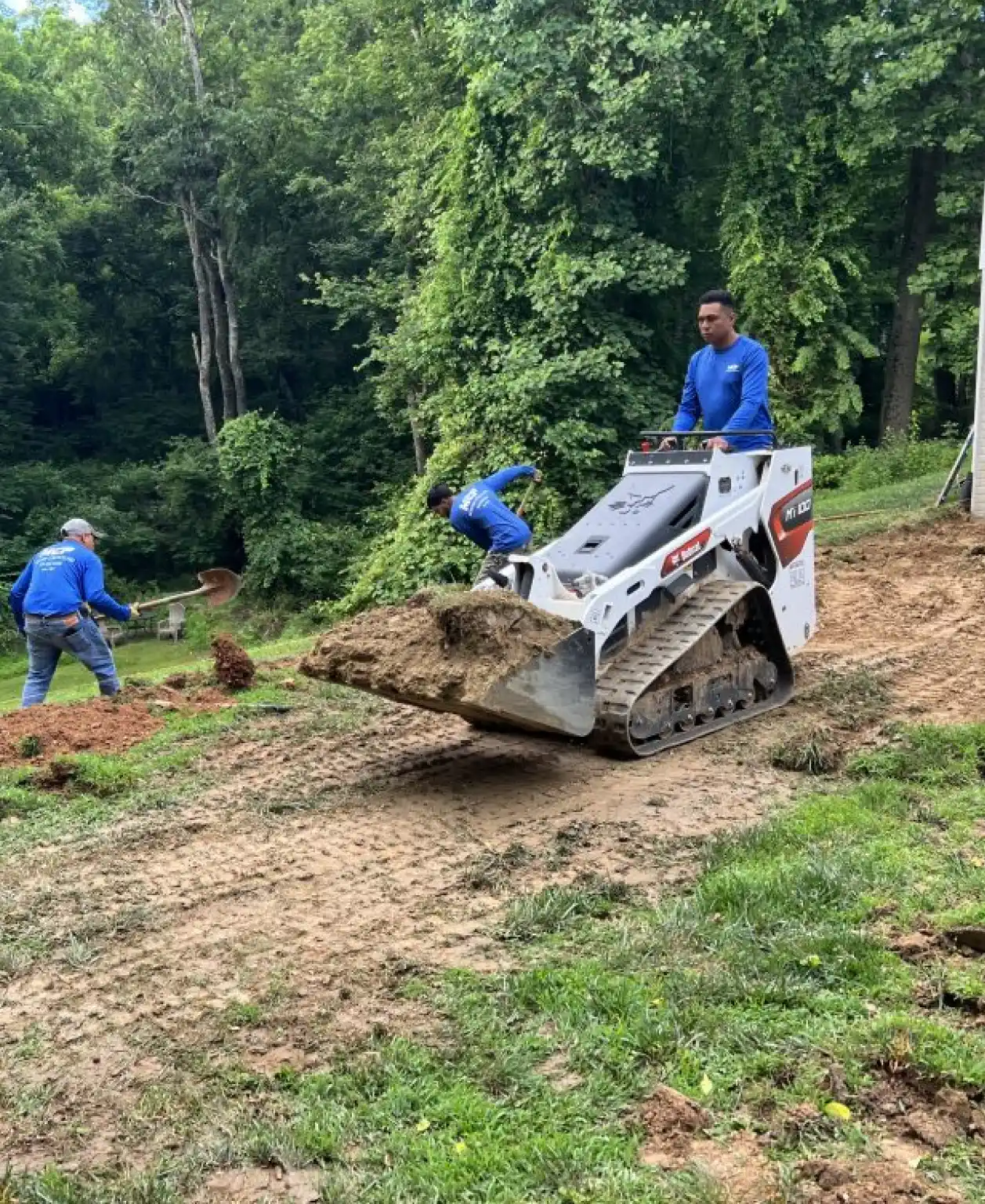 Three workers landscaping; one drives a small track loader carrying dirt, two work with shovels nearby.