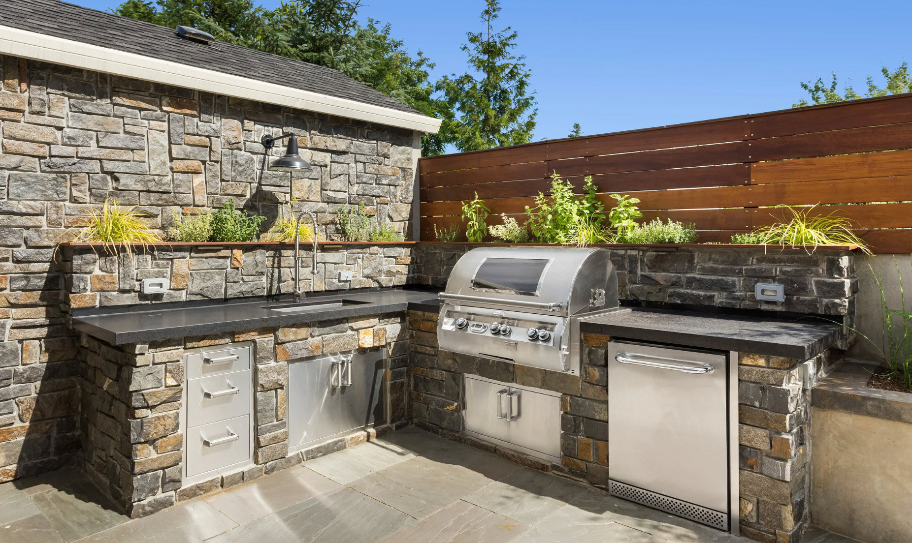 Outdoor kitchen with stone counters, built-in grill, sink, mini fridge, and planters against a wooden fence.