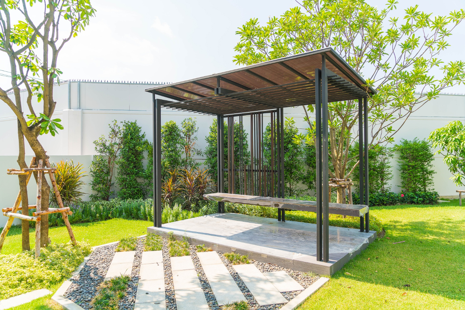 Modern metal and wood pergola with a bench in a landscaped garden with trees and green grass.