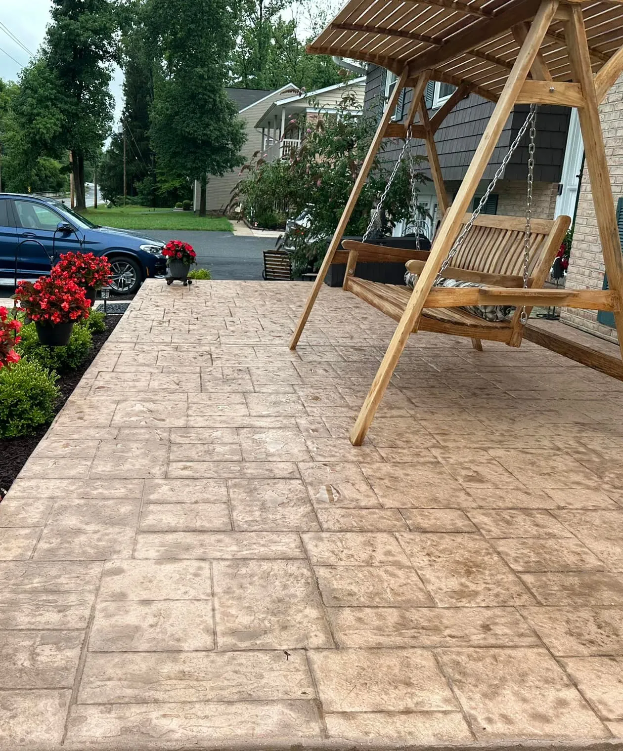Wooden swing on a stamped concrete patio with red flowers and a blue car parked nearby.